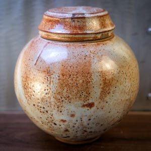 May include: A round ceramic jar with a lid. The jar is primarily off-white with brown speckles and a reddish-brown rim and lid. The jar has a smooth, glossy finish and sits on a wooden surface.