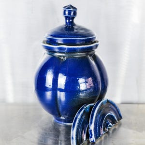 May include: A deep blue ceramic jar with a lid and a scalloped rim. The jar has a rounded body and is decorated with a subtle, textured pattern. The jar is sitting on a silver surface.