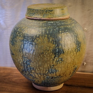 May include: A round ceramic jar with a lid. The jar is primarily blue and yellow with a textured, dripped glaze. The lid is a matching color and shape. The jar sits on a wooden surface.