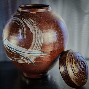 May include: A brown ceramic jar with a lid. The jar has white and black brushstrokes on the surface. The lid is brown with a spiral design.
