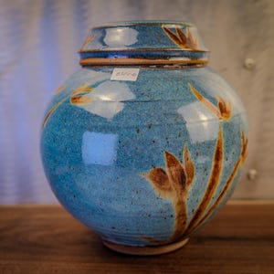 May include: A round, blue ceramic jar with a lid. The jar features brown and orange floral designs. The jar is sitting on a wooden surface. The jar has a label on it.