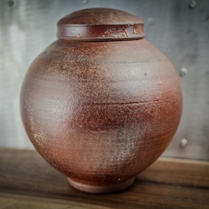 May include: A handcrafted, round ceramic urn with a lid. The urn is a warm, earthy brown color with a textured surface. The lid fits snugly on top. The urn is likely used for decorative purposes or as a container.