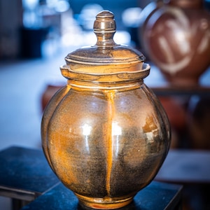 May include: A ceramic jar with a lid, featuring a bulbous body and a dark brown and gold glaze. The jar has a rounded shape with vertical ridges and a small, knob-like handle on the lid. The background shows other similar pottery pieces.