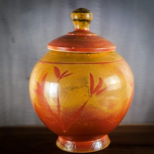 May include: A ceramic jar with a lid, featuring a warm color palette of yellow, orange, and red. The jar has a rounded body and a pedestal base. The lid is topped with a small knob. The jar is decorated with stylized floral motifs.