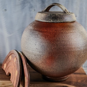 May include: A round, ceramic jar with a lid and handle. The jar is a mottled brown color with a textured surface. The lid is a darker brown with a handle. The jar is sitting on a wooden surface.