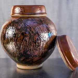 May include: A round, glazed ceramic jar with a lid, featuring a dark brown and black mottled pattern. The lid is off to the side. The jar has a light beige base and a glossy finish, suitable for storage or display.