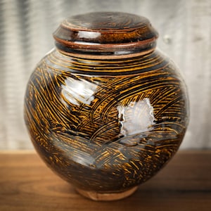 May include: A handcrafted, ceramic urn with a lid. The urn is a deep brown color with a textured, etched pattern in black and gold. The round shape and lid suggest it is designed for storage or display.