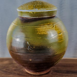 May include: A handcrafted ceramic jar with a lid. The jar is a rounded shape, with a dark brown base that fades into a green and yellow body. The lid is a matching green and yellow with a textured surface. The jar is likely used for storage or decoration.