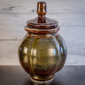 May include: A ceramic jar with a lid, featuring a mottled green and brown glaze. The jar has a rounded body and a slightly flared opening.