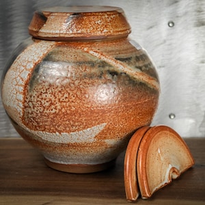 May include: A ceramic jar with a lid, featuring a speckled orange and brown glaze with white accents. The jar has a rounded shape, and the lid is partially removed, revealing the interior. The jar is sitting on a wooden surface.