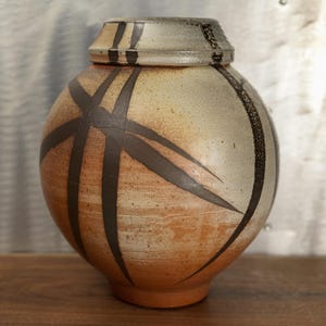 May include: A handcrafted ceramic jar with a lid. The jar is spherical with a tan and orange base, and a cream-colored top. Dark brown lines create a geometric pattern across the surface. The lid has matching dark brown stripes.