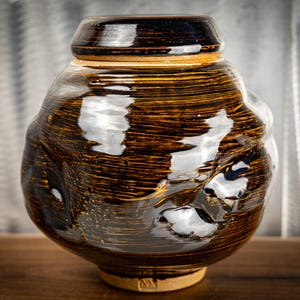 May include: A round ceramic jar with a glossy dark brown glaze featuring horizontal striations and irregular indentations along the body. The vessel has a wide, rounded midsection, a narrow neck, and a matching lid with a dark, reflective finish.