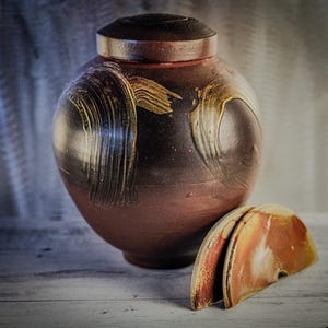 May include: A brown ceramic jar with a lid and a gold and black swirl design. The jar is sitting on a white wooden surface with a matching lid next to it.