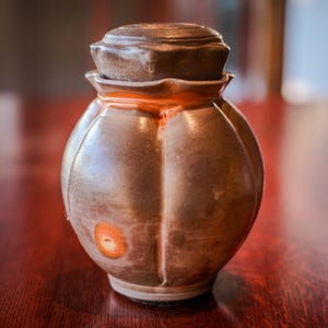 May include: A handcrafted, brown ceramic jar with a lid. The jar has a rounded, segmented body and a textured surface. The lid is a darker shade of brown. An orange-colored accent is visible on the jar's surface. The jar is sitting on a dark wooden surface.