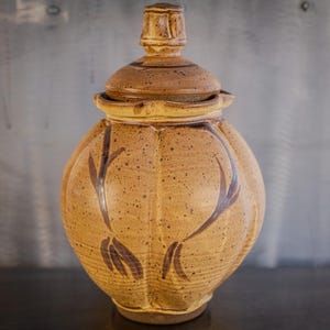May include: A brown ceramic jar with a lid, featuring a speckled glaze and a design of stylized leaves in a darker brown. The jar has a rounded body and a slightly flared opening.