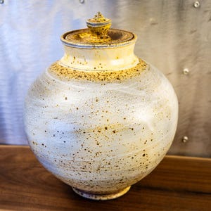 May include: A handcrafted ceramic jar with a lid. The jar has a round body and a cream-colored glaze with brown speckles. The lid and upper portion of the jar are yellow with a textured finish. The jar sits on a wooden surface.