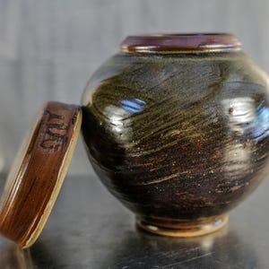 May include: A handcrafted ceramic jar with a lid, featuring a dark, swirling glaze of brown, green, and black. The lid rests beside the round vessel, revealing a glimpse of the interior. The jar's design suggests a focus on pottery and home decor.