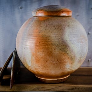 May include: A ceramic urn with a lid, featuring a speckled, tan and orange glaze. The spherical body sits on a base, with a matching lid on top. The urn is displayed on a wooden surface against a textured metal background.