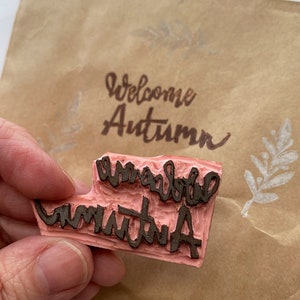 May include: A pink rubber stamp with the words "Welcome Autumn" in a handwritten style. The stamp is held in a hand, with the stamped image visible on a brown paper envelope. The envelope also has a leaf design.