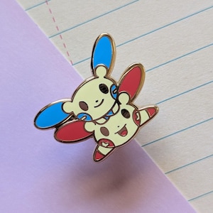 May include: Gold-colored enamel pin featuring two cartoon characters, one yellow with blue ears and the other yellow with red ears, hugging each other.