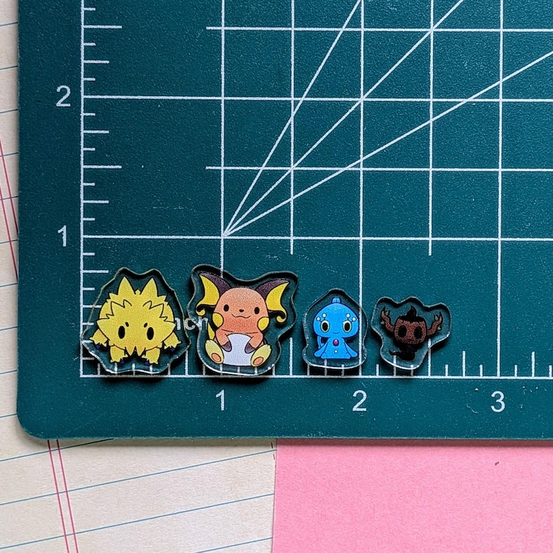 May include: Four small acrylic charms featuring cartoon characters placed on a green cutting mat with white grid lines. The charms include a yellow spiky creature, an orange creature with large ears, a blue round creature, and a dark brown ghost-like figure.