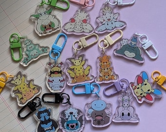 May include: A collection of 14 colorful keychains featuring various cartoon characters. The keychains are made of clear plastic and feature different characters, including a stack of three yellow and orange characters, a stack of three red and orange characters, a blue character with a red hat, and a green character with a red hat. The keychains are all attached to silver metal rings.
