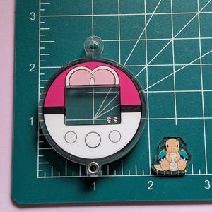 May include: A circular acrylic charm designed like a pink and white Poke Ball with a digital screen interface and a small metal loop at the top and bottom. Next to it is a small, separate acrylic charm featuring a tan dragon character with blue wings.