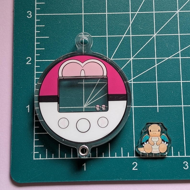 May include: A circular acrylic charm designed like a pink and white Poke Ball with a digital screen interface and a small metal loop at the top and bottom. Next to it is a small, separate acrylic charm featuring a tan dragon character with blue wings.