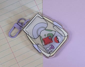 May include: A clear acrylic keychain featuring a cartoon design of a handheld video game console. The keychain has a light purple clasp and a colorful design with red, green, and white accents. The keychain is on a background of lined paper and lavender paper.