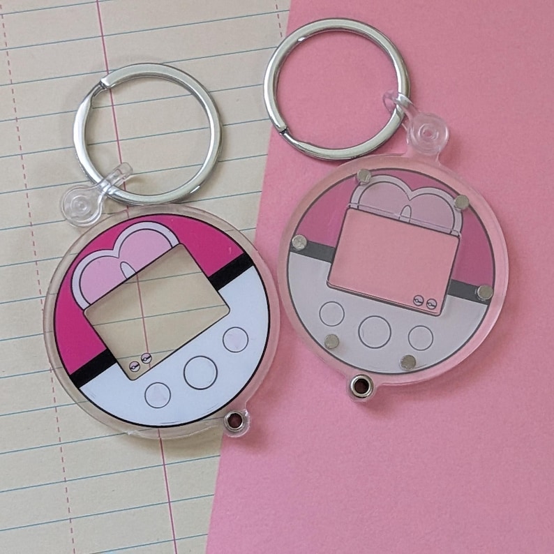 May include: Two circular acrylic keychains featuring a graphic design of a digital pet device. The left keychain is bright pink and white, while the right one is a softer pastel pink and white. Both include silver metal rings for attachment.