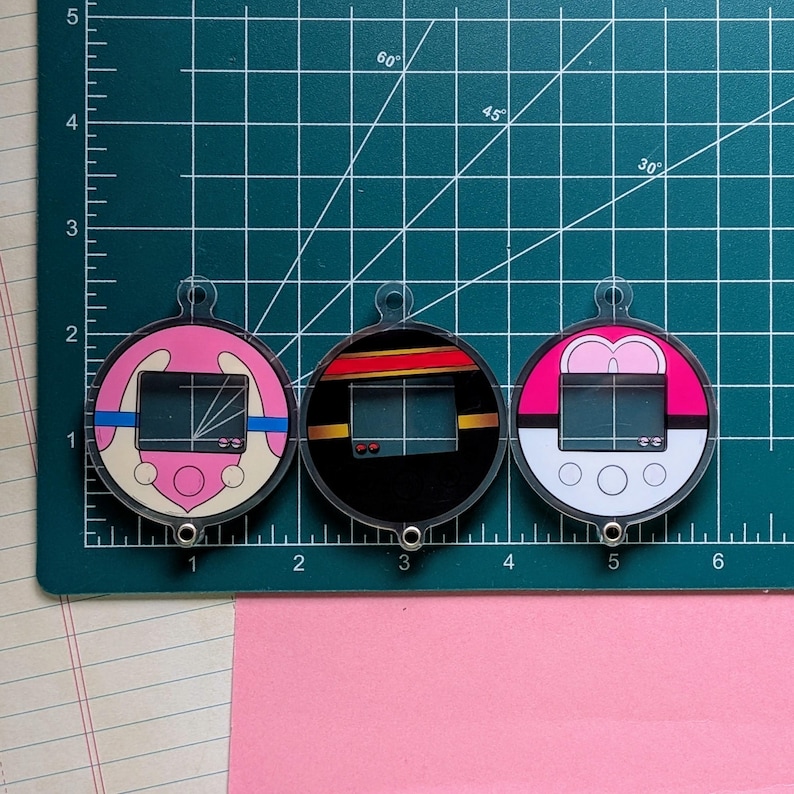 May include: Three circular acrylic charms featuring digital pet toy designs. The left charm is pink and cream, the center is black with red and yellow stripes, and the right is white and pink. Each charm has a small loop and a screen graphic.