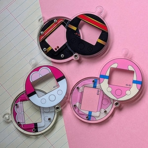 May include: A collection of five circular acrylic charms shaped like digital pet devices. The charms feature various color schemes including black with red and gold stripes, pink and white, and pink with blue accents, each with a central screen cutout.