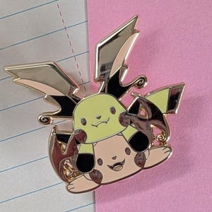 May include: A gold-colored enamel pin featuring a cartoon character with a yellow and tan body, black accents, and gold wings. The pin is set against a pink and lined paper background.