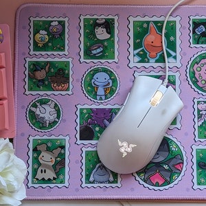 May include: A white wireless gaming mouse with a Razer logo on a pink and green mousepad with cartoon characters. The mousepad has a design of postage stamps with different characters on each stamp.