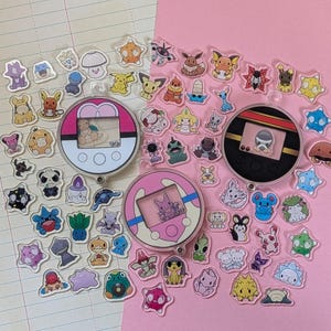 May include: A collection of small, clear acrylic charms featuring various cartoon creatures and three circular, handheld digital pet-style devices. The charms are scattered across a surface divided between lined paper and a solid pink background.