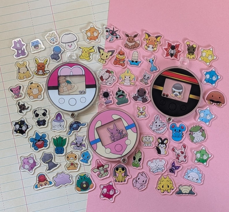 May include: A collection of small, clear acrylic charms featuring various cartoon creatures and three circular, handheld digital pet-style devices. The charms are scattered across a surface divided between lined paper and a solid pink background.