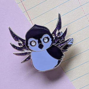 May include: A purple and blue enamel pin of a penguin with wings. The penguin has large, white eyes and a black beak. The wings are detailed with a purple glitter finish.