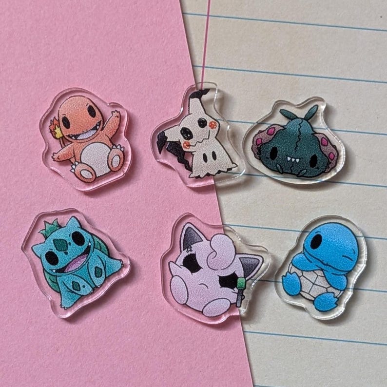 May include: Six small, clear acrylic charms featuring colorful cartoon character illustrations. The set includes an orange lizard, a cream-colored ghost, a dark green creature, a teal plant-based creature, a light pink round character, and a blue turtle.