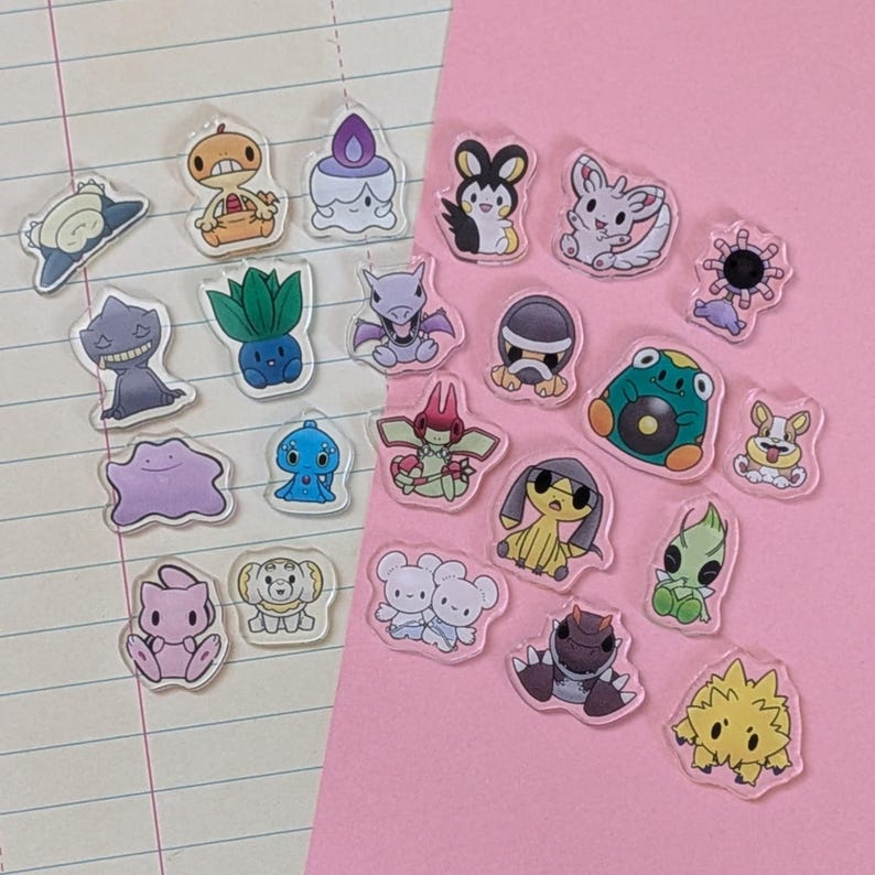 May include: A collection of twenty-two small, clear acrylic charms featuring colorful cartoon character illustrations. The charms depict various creatures in diverse poses and colors, including shades of purple, green, yellow, and blue, arranged on a split background of lined paper and solid pink.