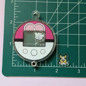 May include: A circular acrylic charm shaped like a pink and white ball with a screen displaying two small white creatures and a separate small acrylic charm of a tan and white dog, both placed on a green cutting mat with imperial inch markings.