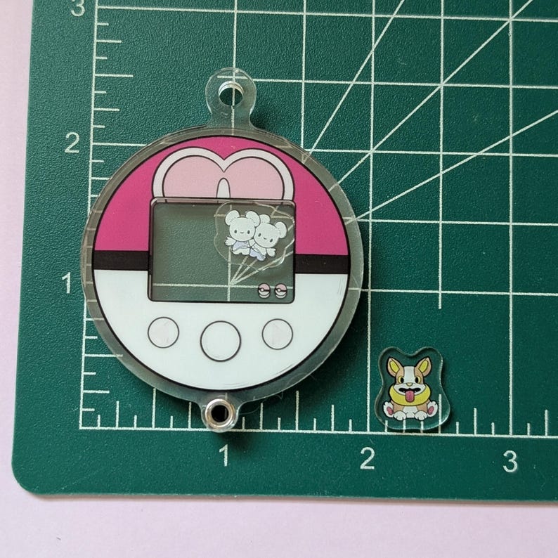 May include: A circular acrylic charm shaped like a pink and white ball with a screen displaying two small white creatures and a separate small acrylic charm of a tan and white dog, both placed on a green cutting mat with imperial inch markings.