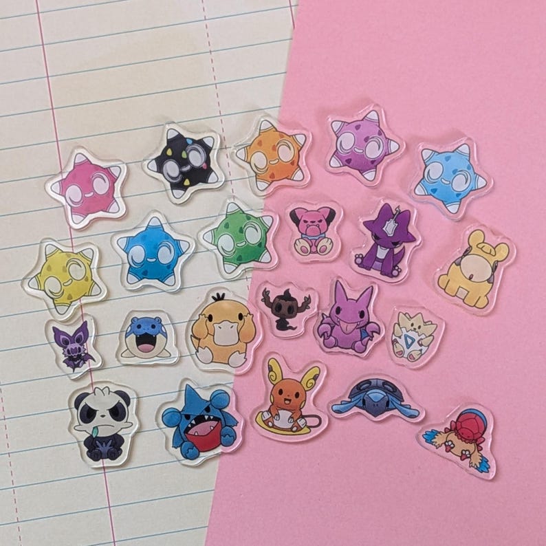 May include: A collection of small, clear acrylic charms featuring various colorful cartoon creatures. The assortment includes star-shaped designs with faces, small animal-like figures, and diverse characters in shades of pink, blue, yellow, and purple.