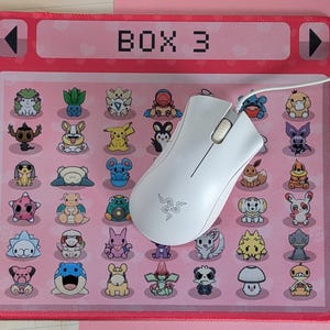 May include: A pink rectangular mousepad featuring a grid of colorful cartoon characters and the text "BOX 3". A white computer mouse rests on the pad, with a white cord extending from it.