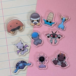 May include: A collection of ten clear acrylic charms featuring colorful cartoon creatures. The set includes a brown dome-shaped figure, a gray turtle-like character, a blue long-necked creature, a purple winged figure, and various other stylized animal designs.
