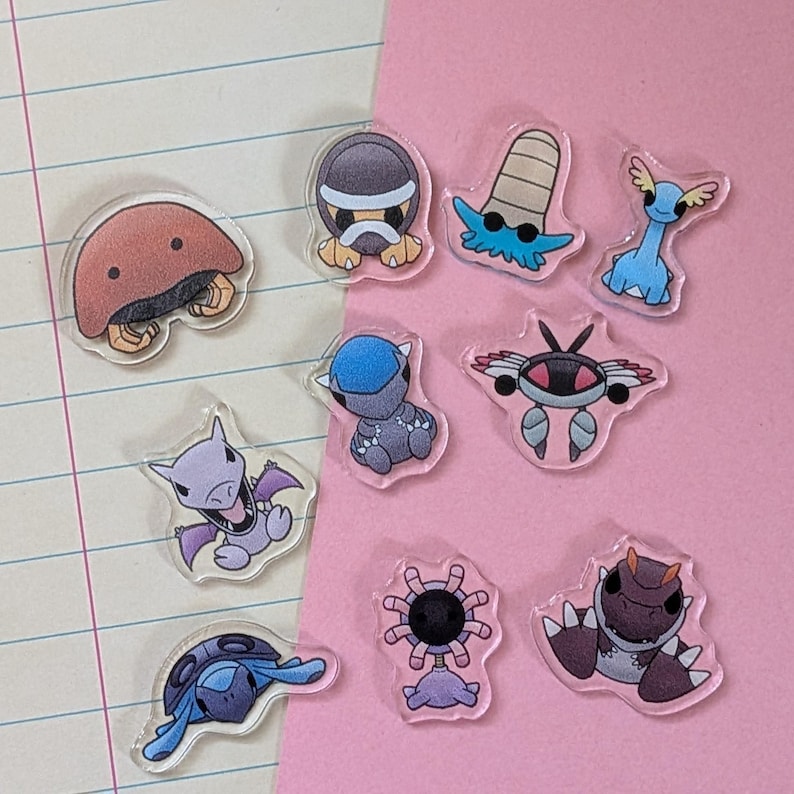 May include: A collection of ten clear acrylic charms featuring colorful cartoon creatures. The set includes a brown dome-shaped figure, a gray turtle-like character, a blue long-necked creature, a purple winged figure, and various other stylized animal designs.