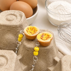 Könnte beinhalten: Ein Paar Ohrhänger mit Miniatur-Eiertörtchen-Anhängern, die goldbraune Krusten und karamellisierte gelbe Füllungen aufweisen, ergänzt durch zwei kleine gelbe Perlen an silberfarbenen Metallhaken für Ohrlöcher.
