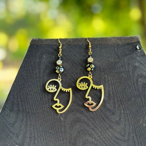 May include: Gold earrings with a face outline design and black and gold beads. The earrings are hanging from a black surface.