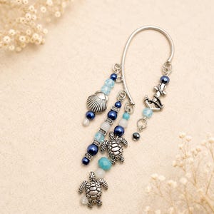 May include: A silver-toned ear cuff featuring dangling charms including two sea turtles, a seashell, and an anchor. The design incorporates various beads in shades of blue, white, and turquoise, attached to the metal frame with small silver jump rings.