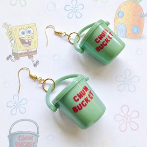 Könnte beinhalten: Ein Paar Ohrhänger in Form von hellgrünen Miniatur-Eimern mit dem in Rot aufgedruckten Text CHUM BUCKET. Jeder Eimer verfügt über einen goldfarbenen Metallhaken und einen kleinen kreisförmigen Ring zur Befestigung am Ohrläppchen.