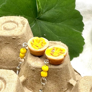 May include: Handmade earrings featuring miniature, detailed egg tarts. The earrings have a yellow and brown color scheme, with yellow beads and silver-colored hooks. The egg tarts are set against a neutral background.